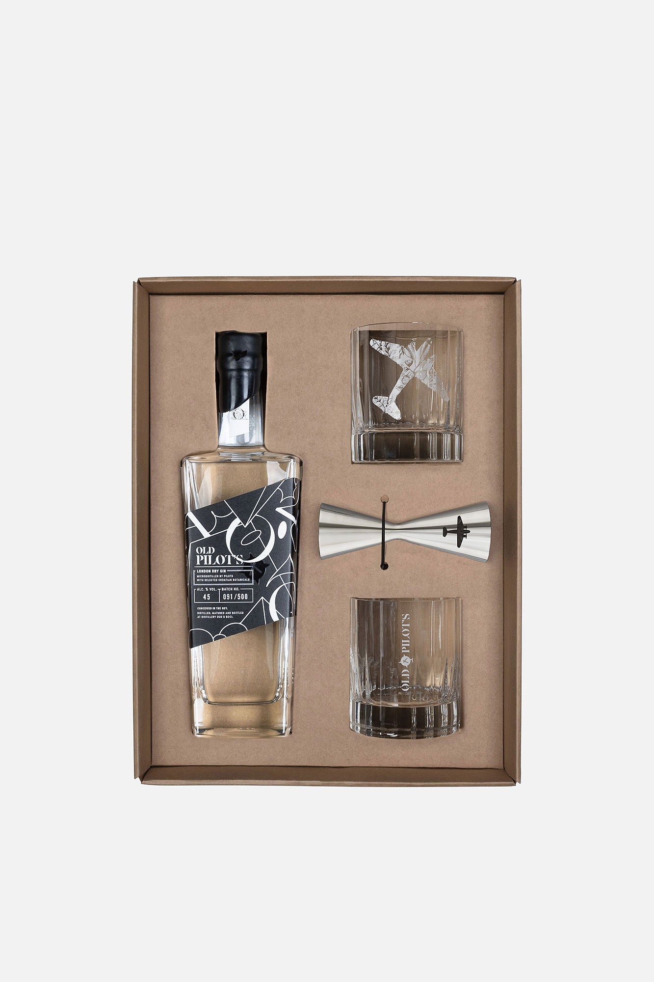 A boxed gift set containing a clear glass bottle with a black cap and dark label, two clear drinking glasses, and a silver jigger, arranged inside a brown presentation box on a light gray background.