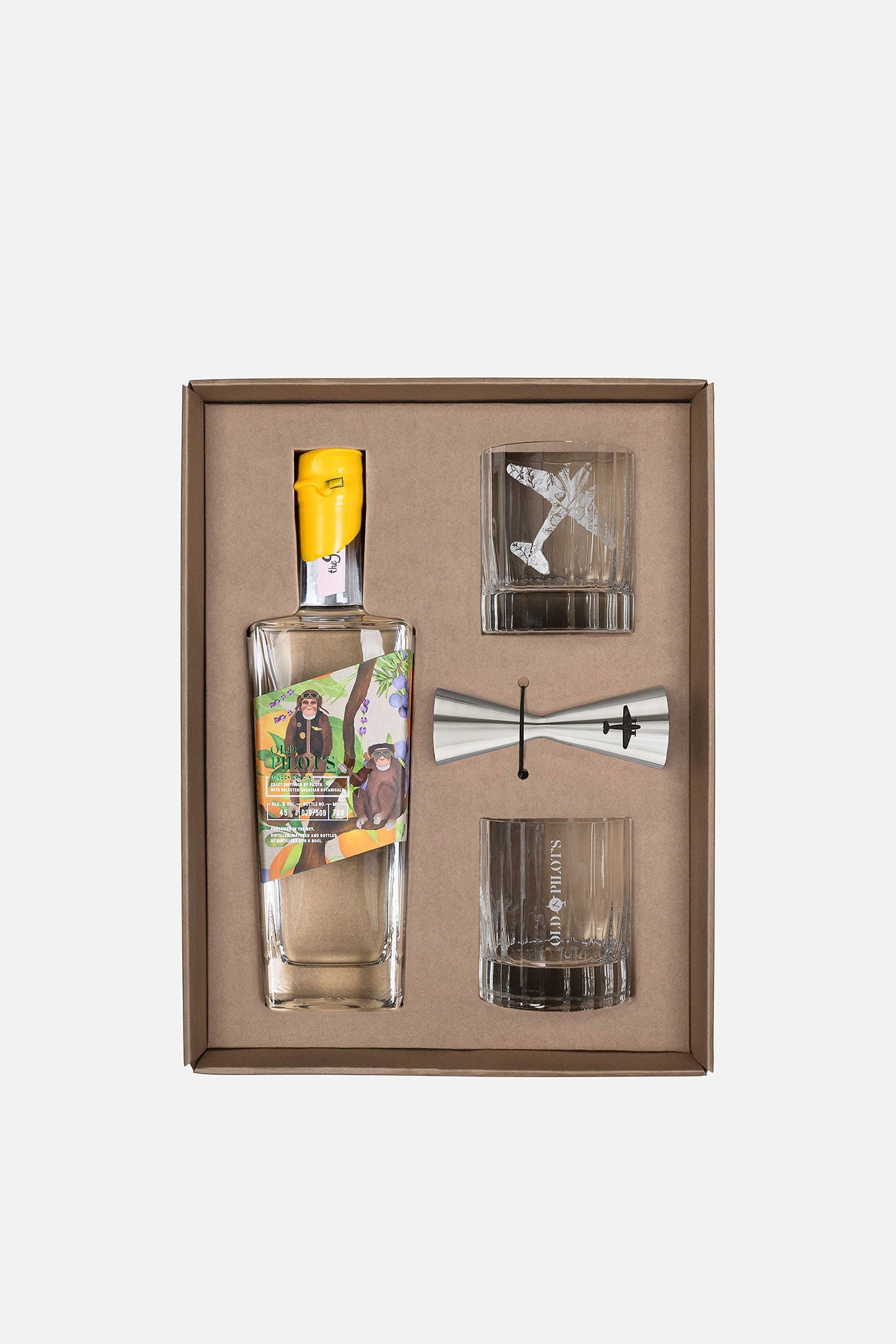 A boxed gift set containing a clear glass bottle with a yellow cap and colorful printed label, plus two clear glasses and a silver jigger, arranged in a brown presentation box on a light gray background.