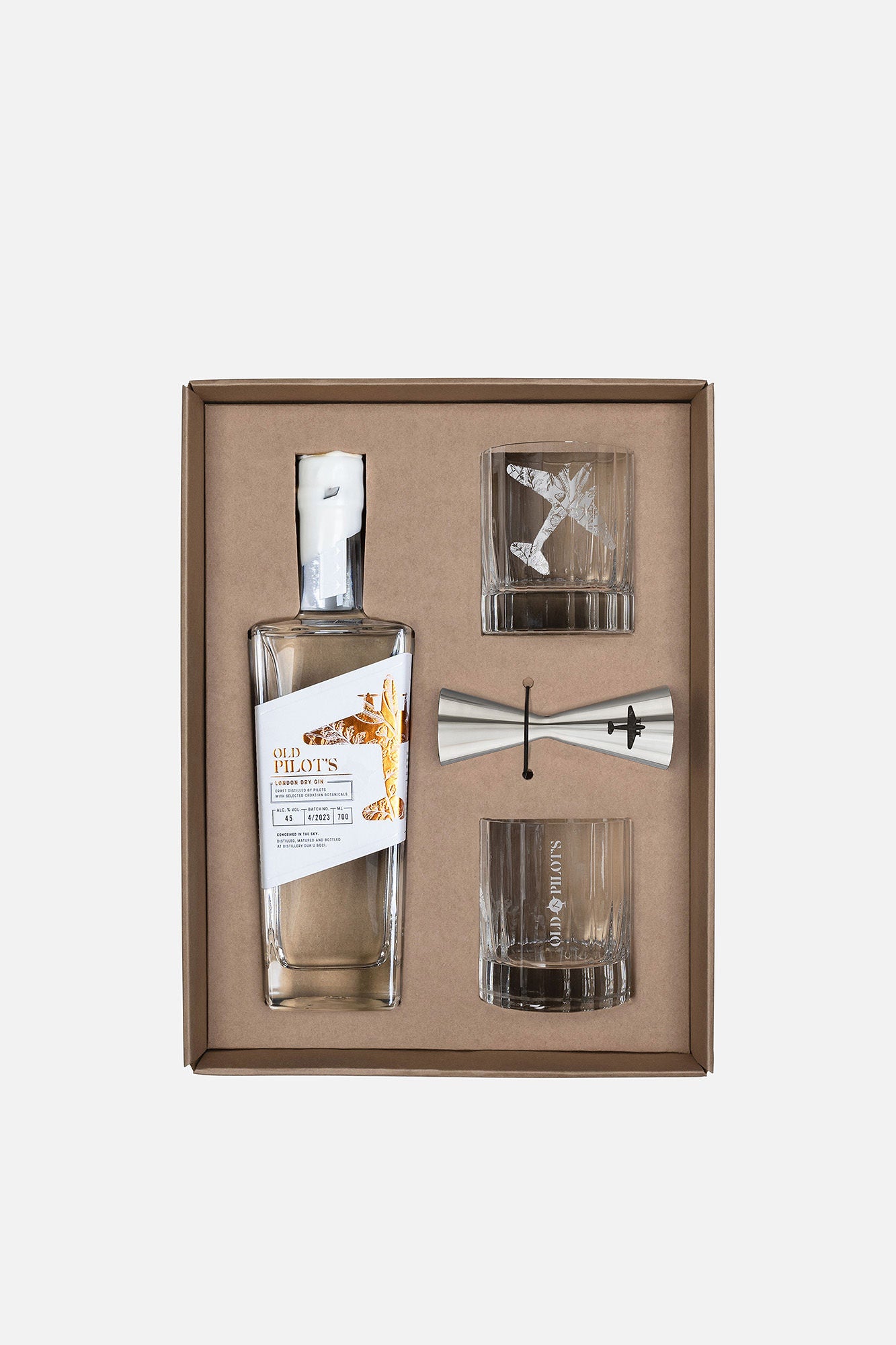 A gift set in a brown box containing a bottle of The Pilot's Gin, a double jigger, and two engraved crystal tumblers on a light gray background.