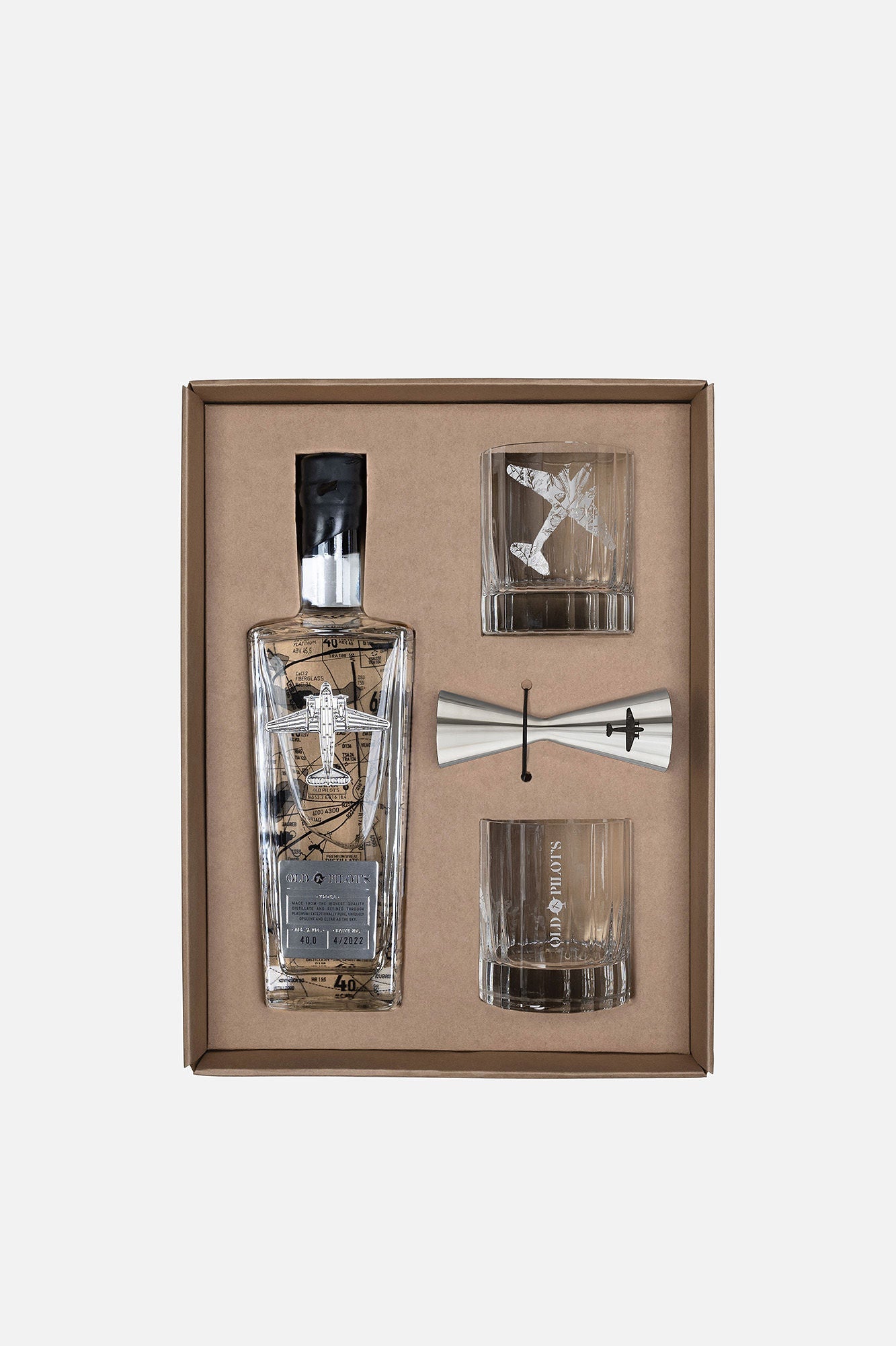 A premium gift set in a brown box containing a clear glass bottle with a black cap, two clear glasses, and a silver jigger, all arranged on a light gray background.